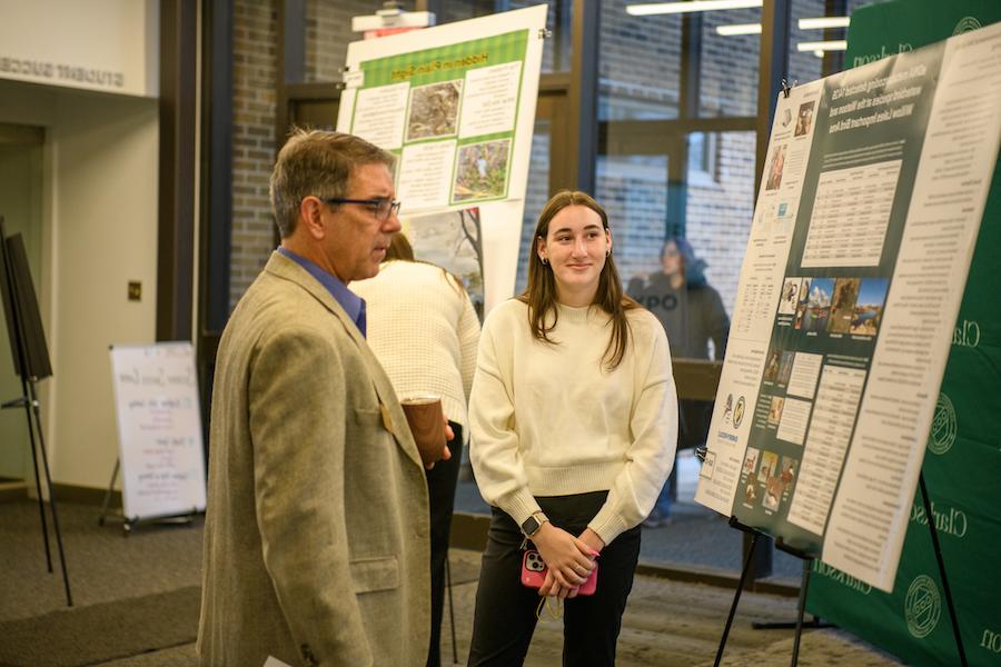 A female student smiles while a male professor reads her poster board.