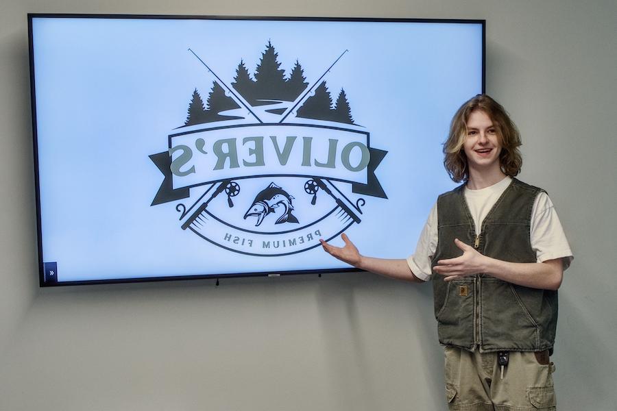 A male student smiles while pitching for a business, with the words “Oliver’s Premium Fish” shown on an electronic screen.